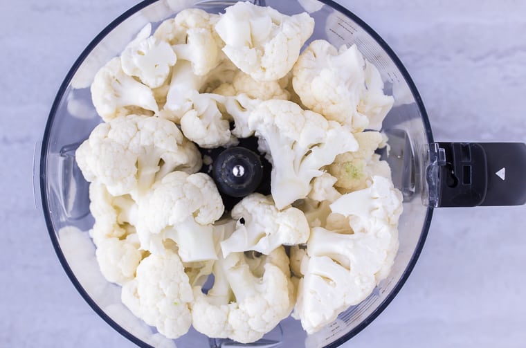 cauliflower florets in the bowl of a food processor over a white background