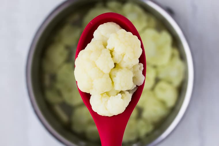 A red spoon filled with boiled cauliflower being held up over the silver pot of cauliflower florets