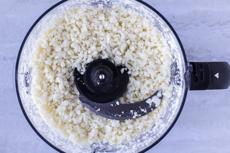 Cauliflower Rice in the bowl of a food processor fitted with a blade over a white background