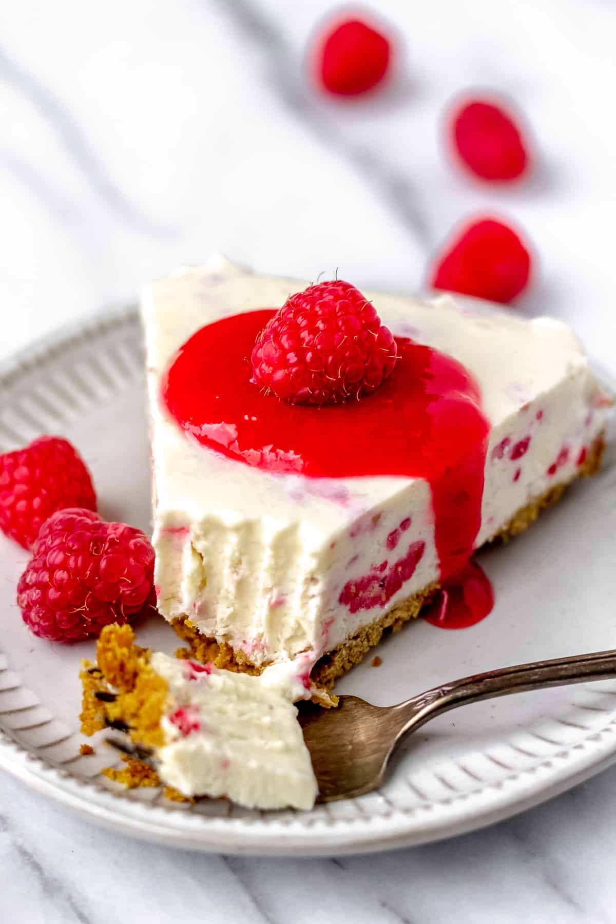 A fork of raspberry cheesecake sitting in front of the rest of the slice on a small plate with raspberries in the background.