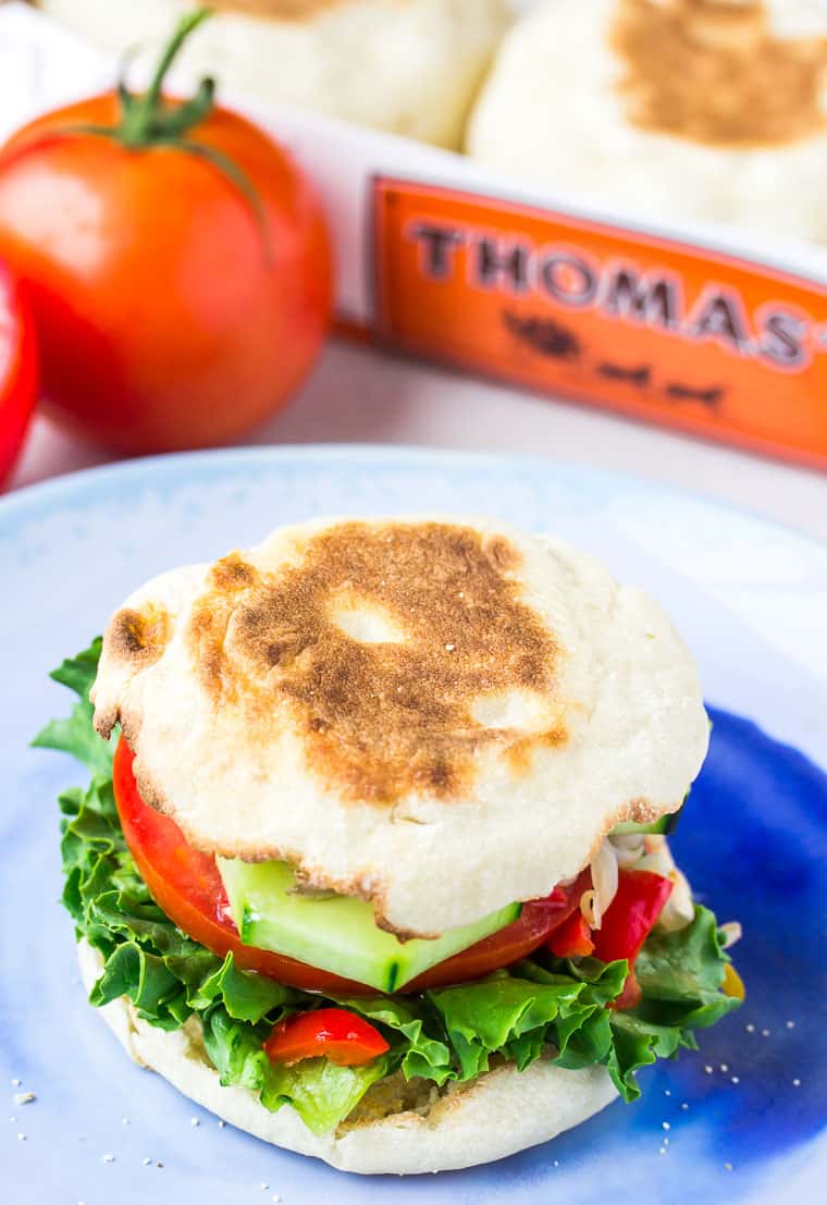 Veggie Sandwich on an English Muffin on a blue plate with tomatoes and thomas' english muffin pack in the background