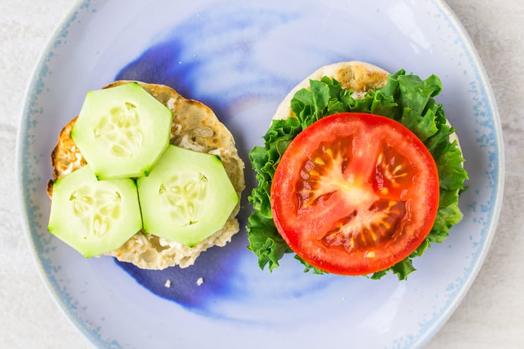 English muffin halves topped with lettuce, tomato, and cucumber on a blue plate over a white background