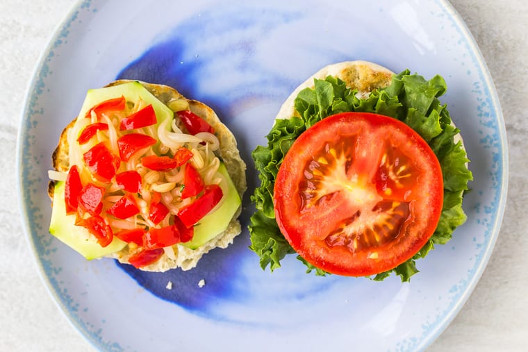 English muffin halves topped with lettuce, tomato, sprouts, red peppers and cucumber on a blue plate over a white background