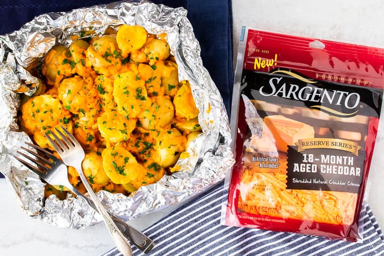 Grilled Cheesy Potatoes in foil next to a package of sargento cheese with a blue napkin and a black and white striped napkin over a white background