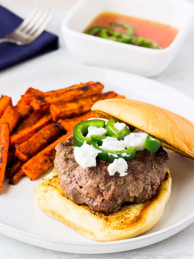 Blue Apron's Jalapeno Burger topped with goat cheese and peppers with a side of carrots, a blue napkin, fork, and pickled jalapenos in the background