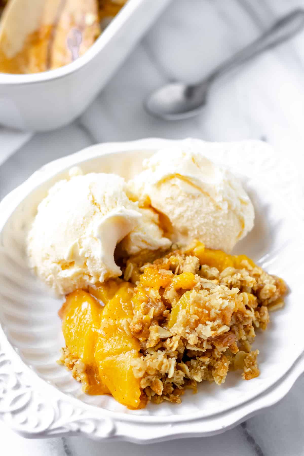 Close up of a serving of peach crisp in a white bowl with ice cream and a spoon and part of the baking dish in the background