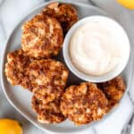 A white plate with shrimp cakes and a bowl of aioli on it with lemons around it.