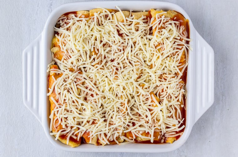 Enchiladas topped with sauce and cheese in a white casserole dish over a white background
