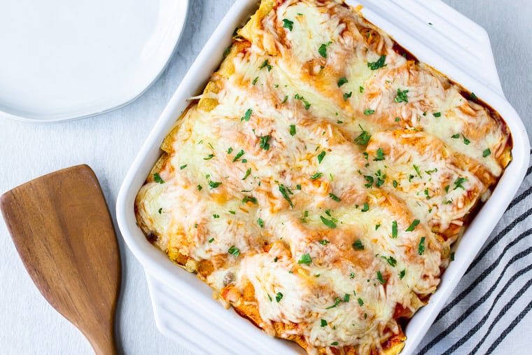 Baked Ground Beef and Mushroom Enchiladas in a white casserole dish on a white background with a blue and white striped towel, and a wood spatula 
