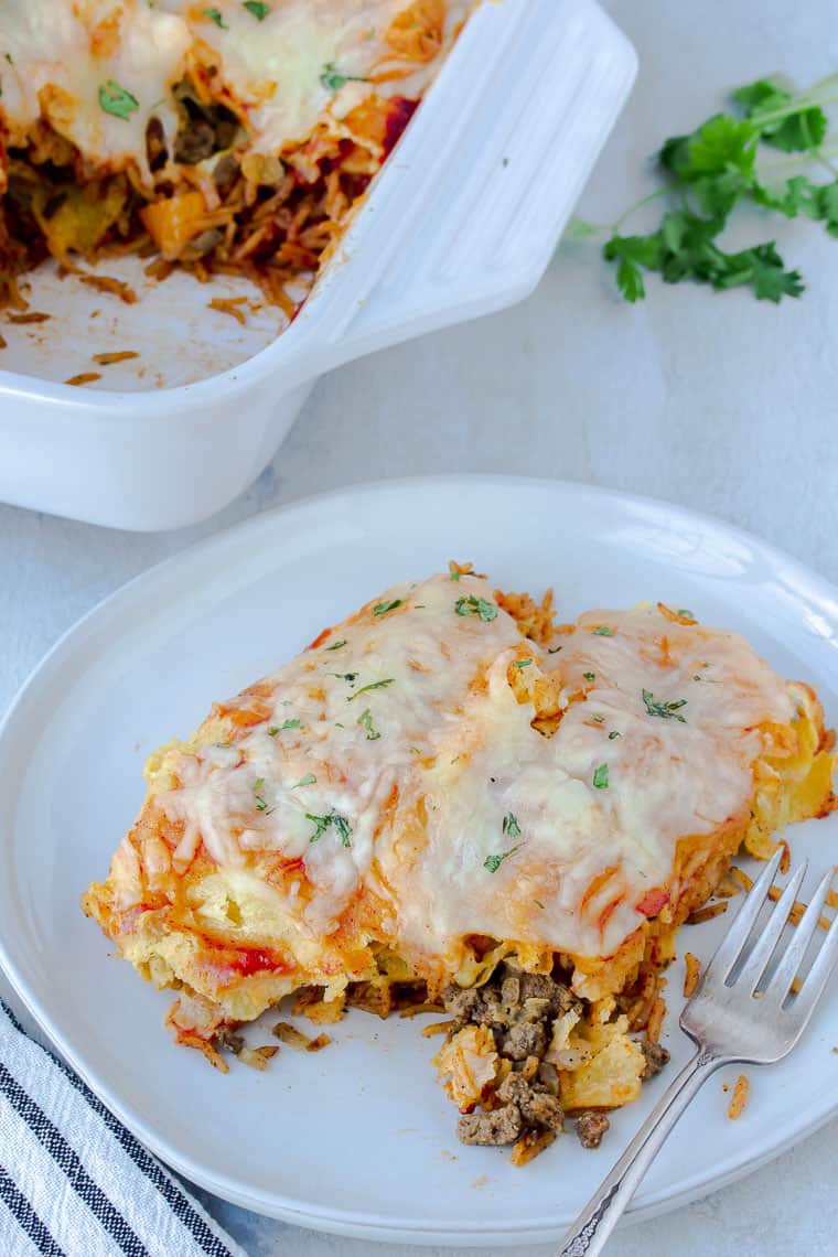 Ground beef and mushroom enchiladas on a plate, cut open with a casserole dish, blue and white striped towel, and cilantro in the background.