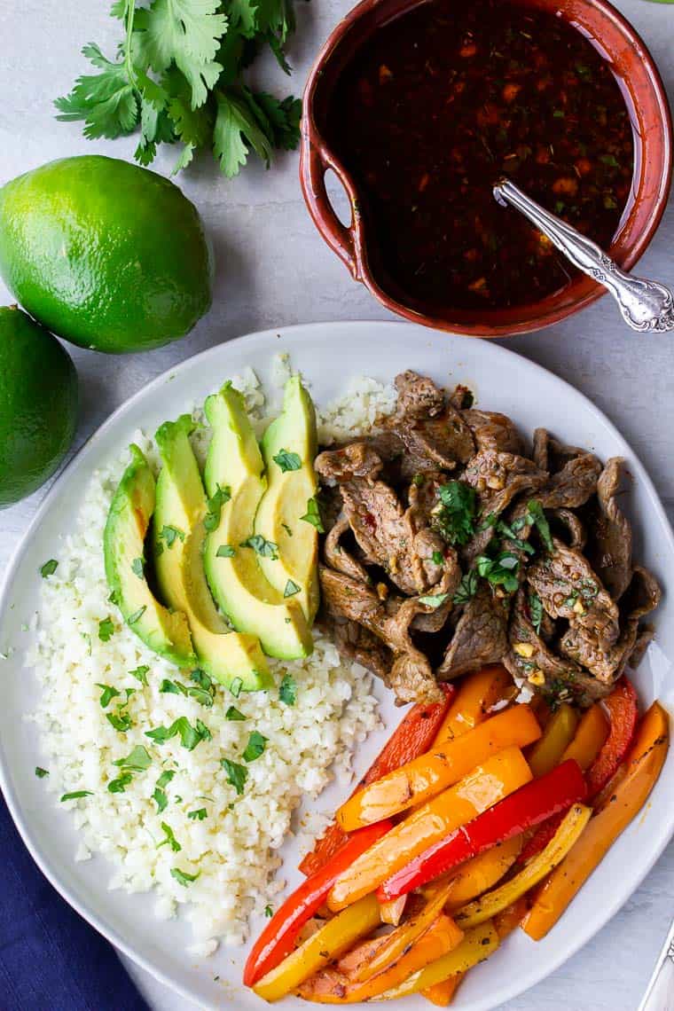 Keto beef fajitas bowls on a white background with an orange bowl of sauce, 2 limes and cilantro