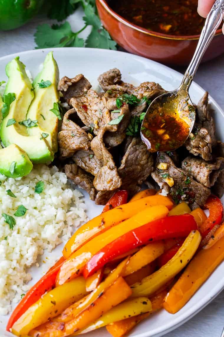 Sauce being spooned over a plate of beef fajitas with peppers, cauliflower rice, and avocado slices