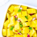 Overhead close up of spicy mango salsa in a white square bowl over a white background with a white napkin