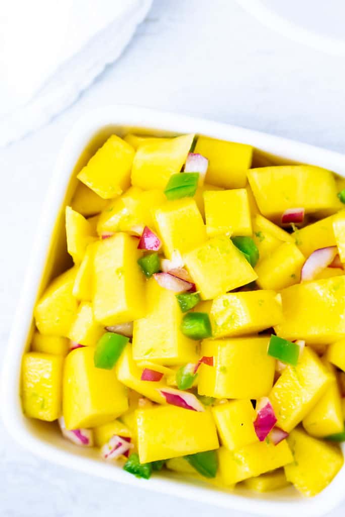 Overhead close up of spicy mango salsa in a white square bowl over a white background with a white napkin
