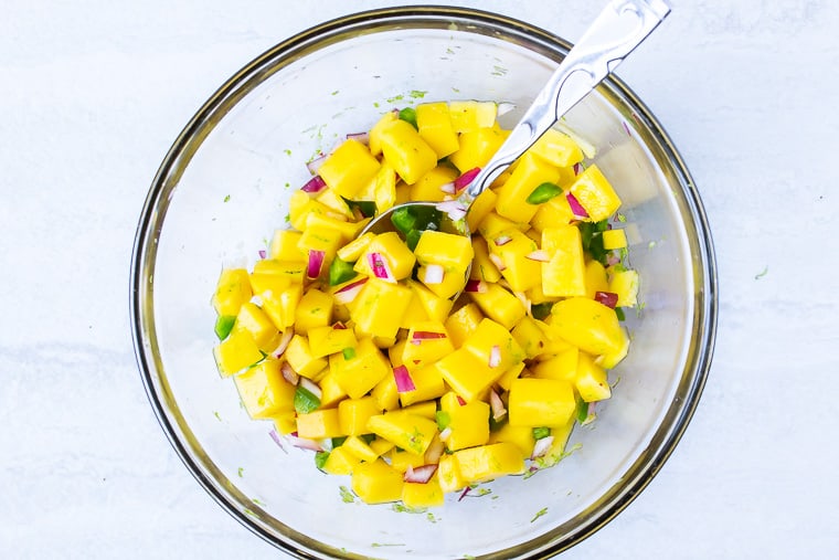 Diced mango, jalapeno peppers, red onion, lime juice and cilantro in a glass bowl with a spoon over a white background
