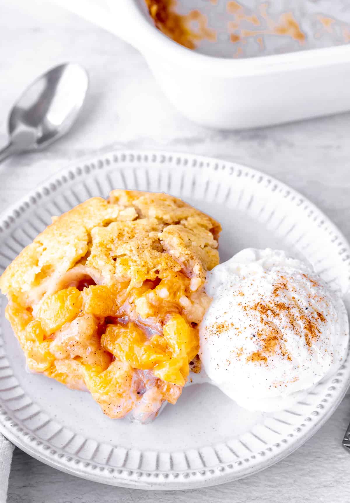 A serving of peach cobbler with vanilla ice cream and the baking dish and a spoon in the background