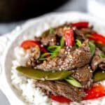 Close up of ginger beef stir fry with sugar snap peas and red peppers in a white bowl with rice with a skillet partially showing in the background with text overlay.