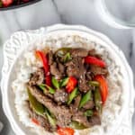 Overhead view of ginger beef stir fry with sugar snap peas and red peppers in a white bowl with rice with a skillet and glass of water partially showing in the background with text overlay.