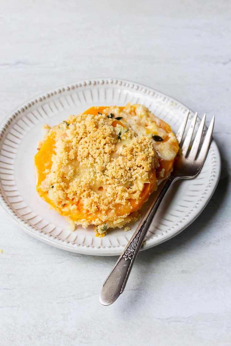 A serving of Butternut Squash Gratin on a small cream colored plate with a fork over a white background