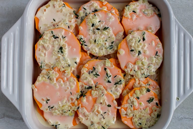 Slices of butternut squash in a white, square casserole dish topped with a cream sauce and herbs