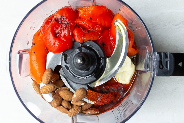 Peppers, almonds, and seasonings in the bowl of a food processor with a blade over a white background
