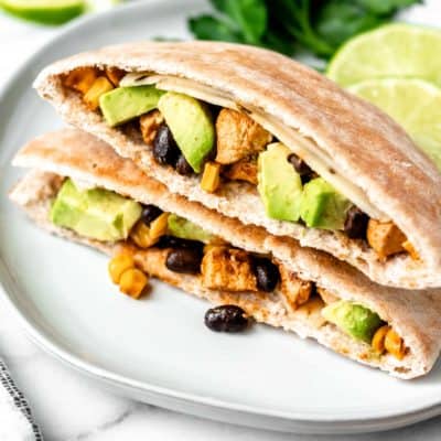 Two Southwestern Chicken Pitas stacked on top of each other on a white plate with cilantro, lime and avocado in the background.