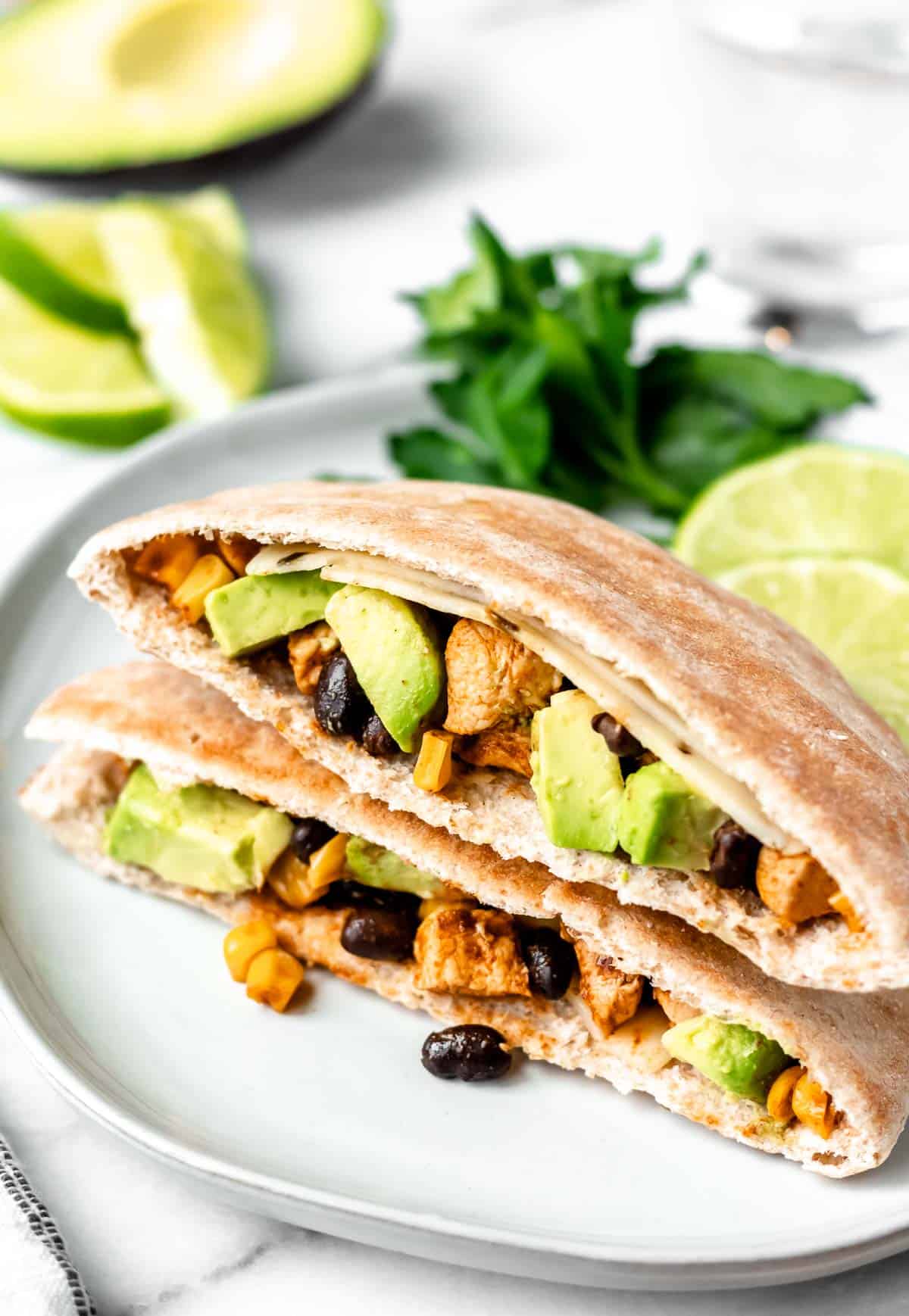 Two Southwestern Chicken Pitas stacked on top of each other on a white plate with cilantro, lime and avocado in the background.