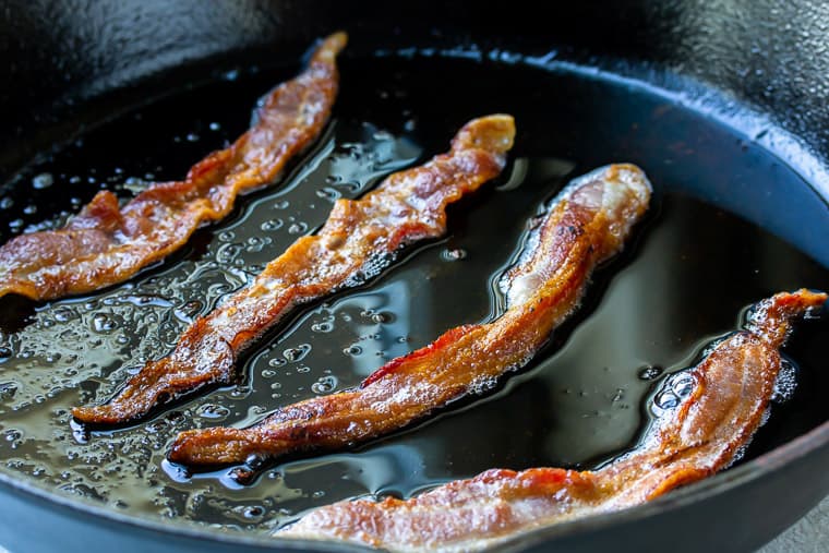 4 slices of bacon cooking in a black cast iron skillet