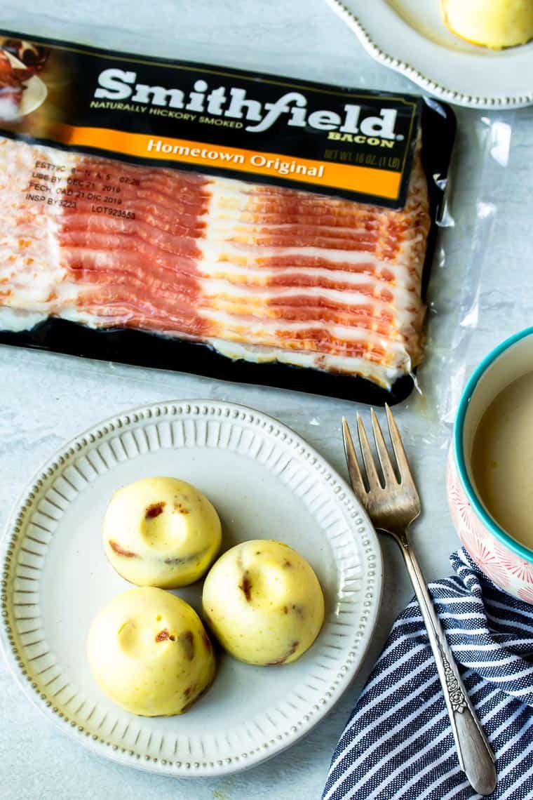 3 Instant Pot Egg Bites on a white plate with a fork, cup of tea, striped napkin, and package of bacon in the background all over a white backdrop