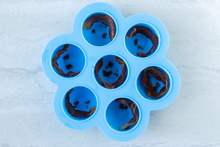 A blue egg bite mold with a piece of bacon in each well over a white backdrop