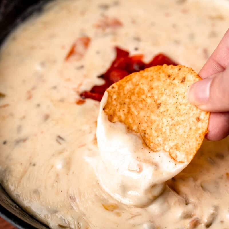A tortilla chip being dipped into a cast iron skillet filled with queso dip.