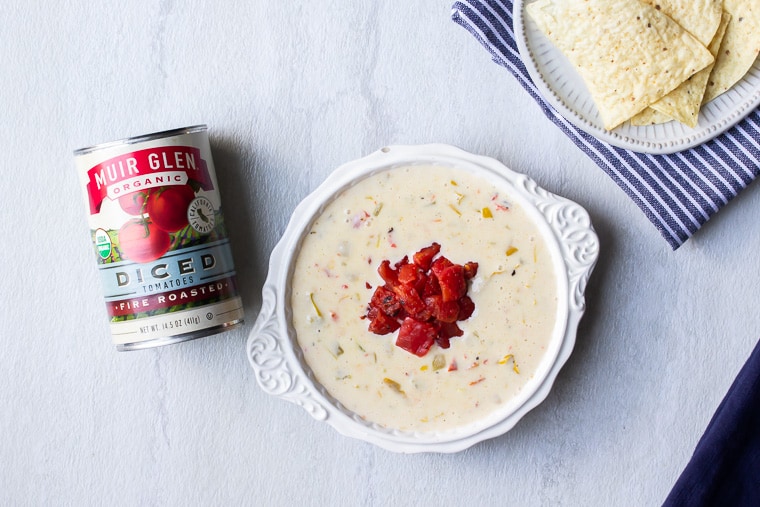 White Queso Dip next to a can of Muir Glen Organic Diced Tomatoes with a blue/white striped napkin and plate of chips next to it