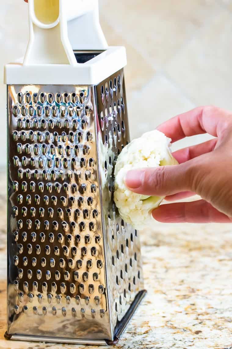 Ricing cauliflower with a box grater over a marble counter and tile background