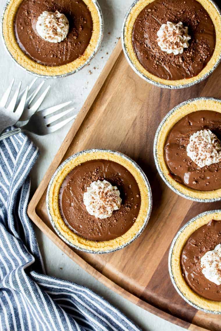4 Mini Chocolate Cream Pies on a cutting board over a white background with another pie and a blue and white striped napkin and 3 forks next to it