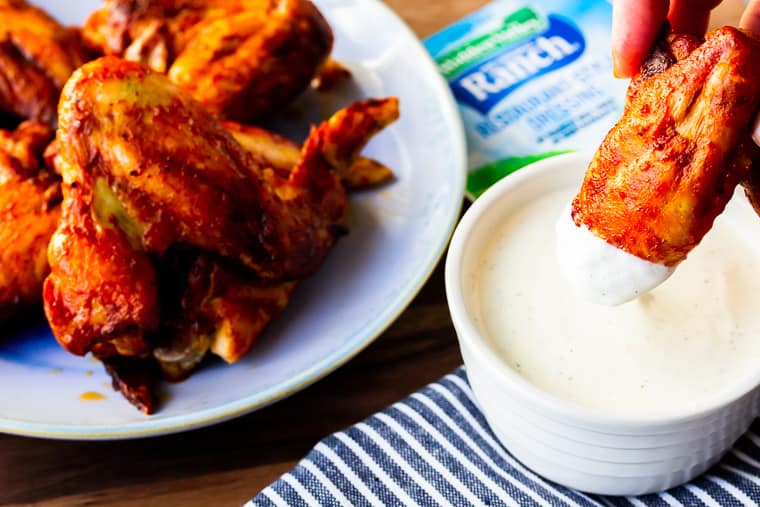 Chicken Wings on a blue plate with one wing being dipped into Ranch Dressing with a blue and white striped napkin and Ranch seasoning mix in the background