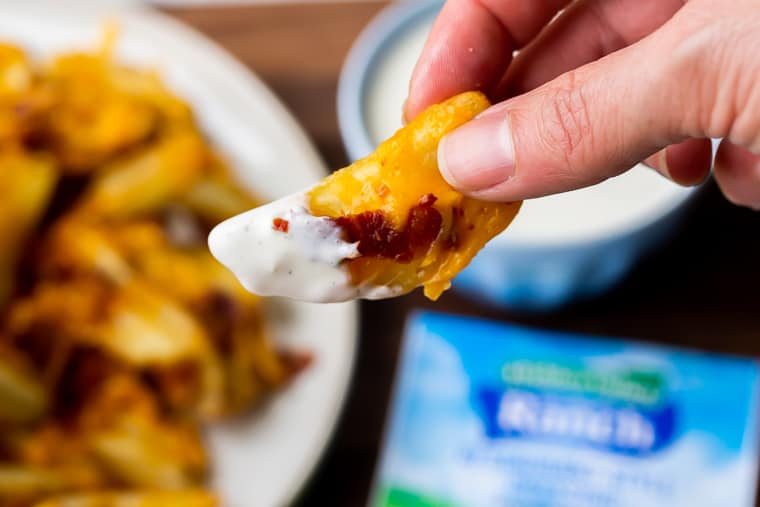 A french fry covered in cheese, bacon, and Ranch dressing being held up with a plate of fries, a packet of Ranch dressing, and a small bowl of Ranch in the background