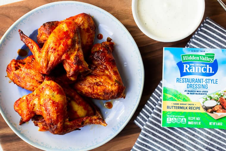 Chicken wings on a blue plate over a wood cutting board with a packet of Ranch, a bowl of Ranch, and a blue and white towel 