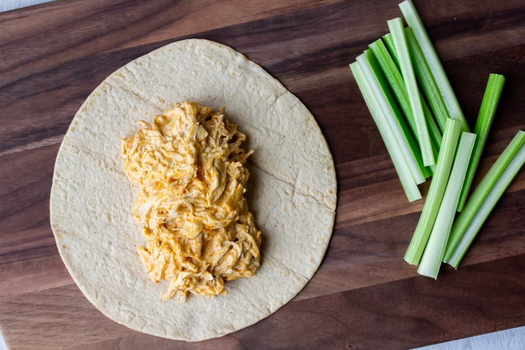 Buffalo chicken on a tortilla with sticks of celery off to the side on a wood cutting board