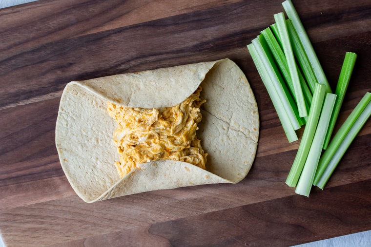 Tortilla filled with buffalo chicken with the top and bottom edges folded over and celery sticks off to the side on a wood cutting board