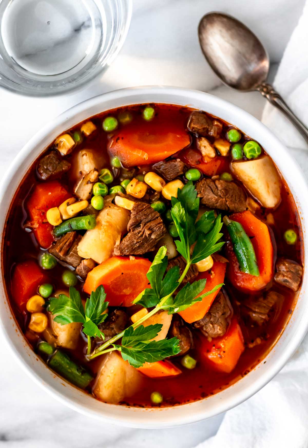 Overhead of Instant Pot Vegetable Beef Soup in a white bowl with a glass of water and spoon around it. g