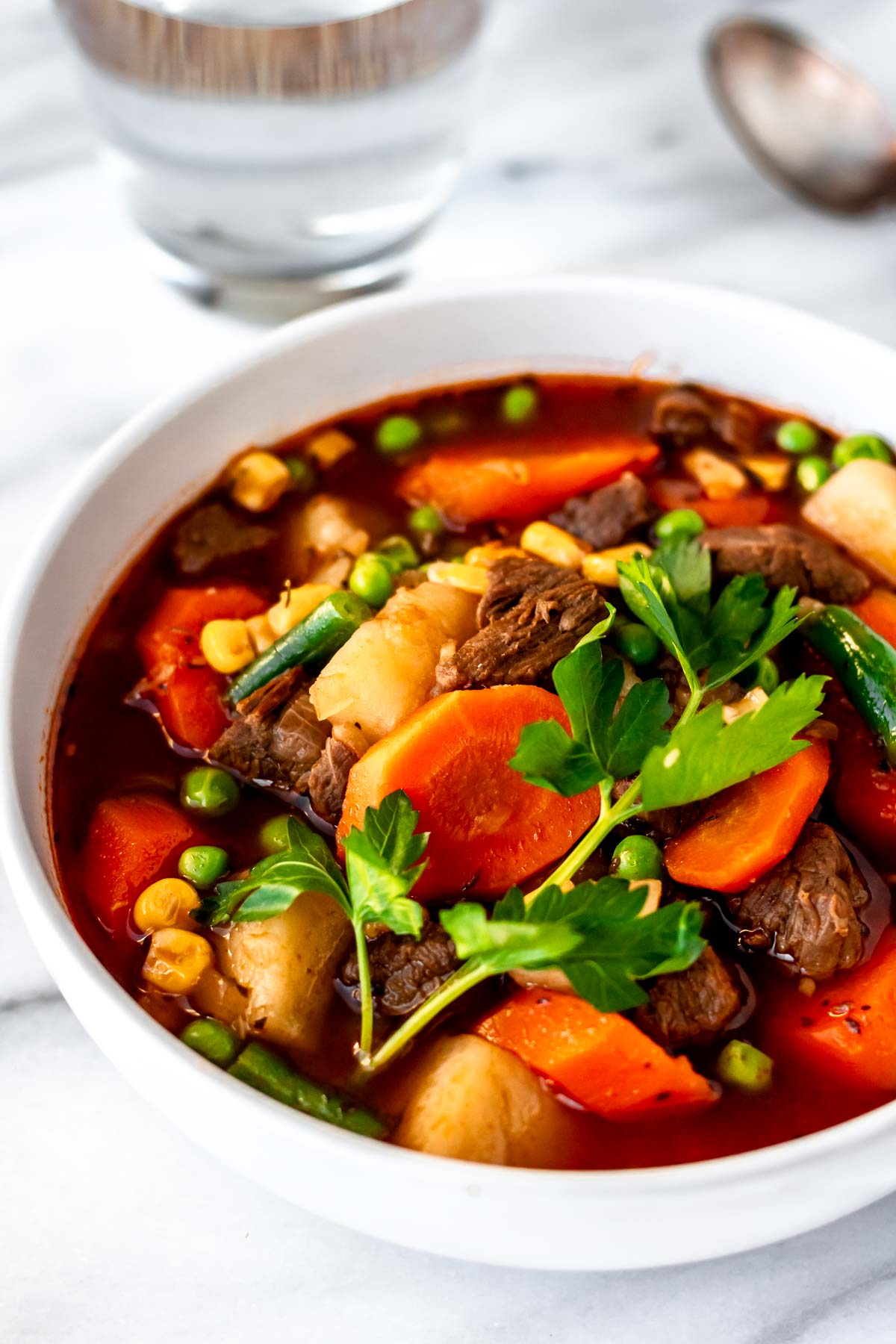 A white bowl with vegetable beef soup in it and a glass of water and spoon in the background.