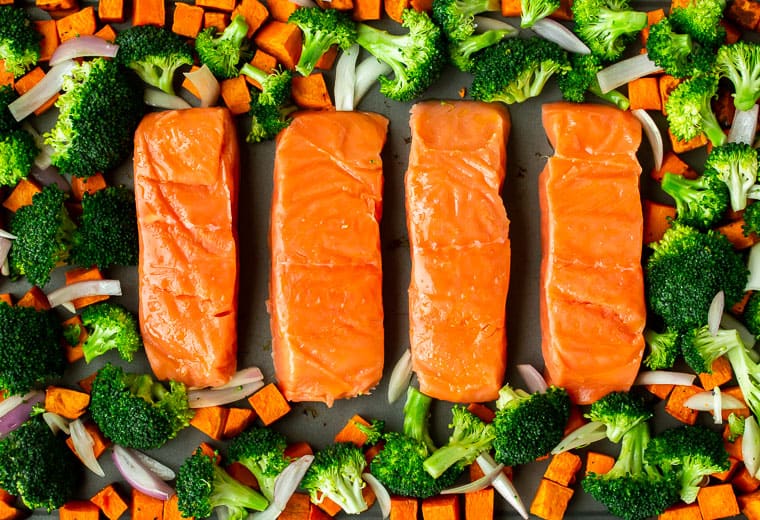 Salmon, broccoli, sweet potatoes. and shallots spread out on a gray sheet pan
