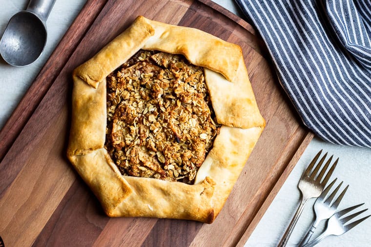 Baked Apple Crostata on a wood cutting board with 3 forks, an ice cream scoop and a blue and white towel in the background
