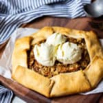 Apple Crisp Crostata topped with 3 scoops of vanilla ice cream on a wood board with a fork, ice cream scoop. and blue and white striped towel in the background
