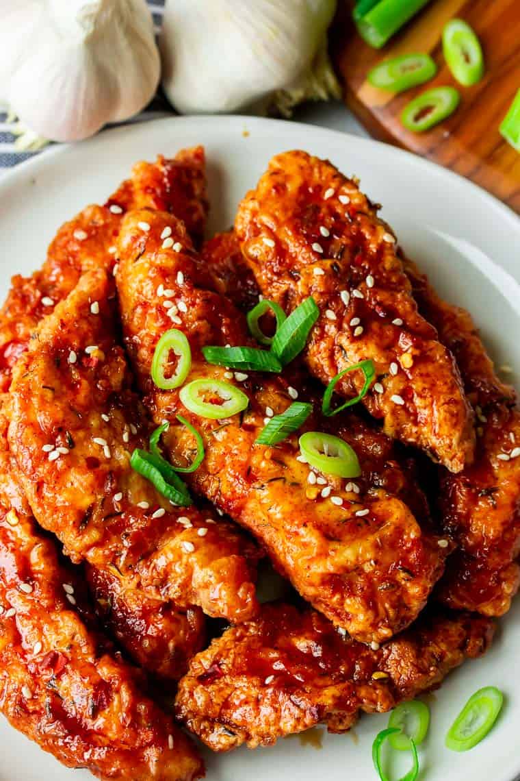 Close up of Honey Garlic Chicken Tenders on a white plate with bulbs of garlic and slices of green onion in the background