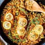 Chicken breasts with lemon slices in a skillet full of orzo with a wood spoon in it. There is a blue an white striped towel under the cast iron pan and part of a bulb of garlic and plate of chicken in the background all over a blue gay background