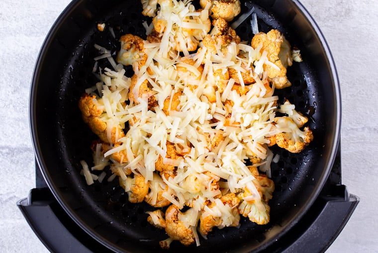 Buffalo cauliflower in an air fryer basket with shredded cheese on top over a white background