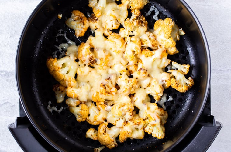 Buffalo cauliflower with melted cheddar cheese on top of it in an air fryer basket over a white background