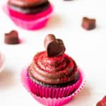 A single chocolate cupcake with chocolate frosting on a white background with a second one blurred in the background and heart shape chocolate candies around it