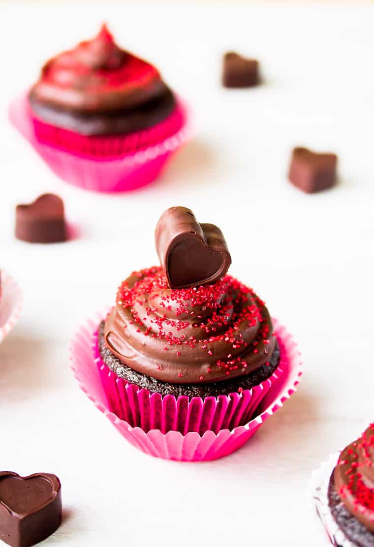 A single chocolate cupcake with chocolate frosting on a white background with a second one blurred in the background and heart shape chocolate candies around it